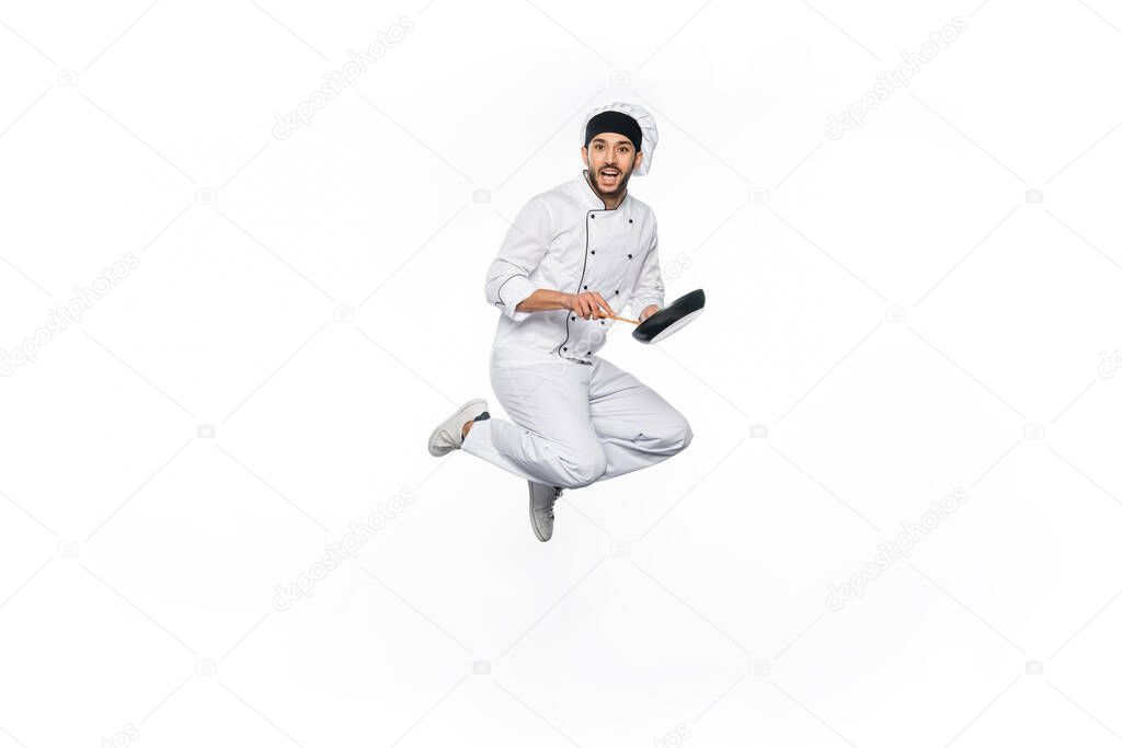 cheerful chef in hat and uniform jumping and holding frying pan with wooden spatula isolated on white 
