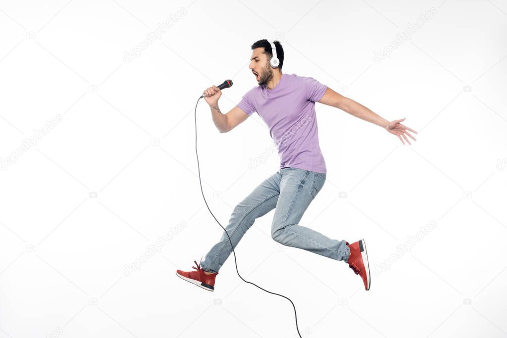 young man in wireless headphones singing in microphone and levitating on white