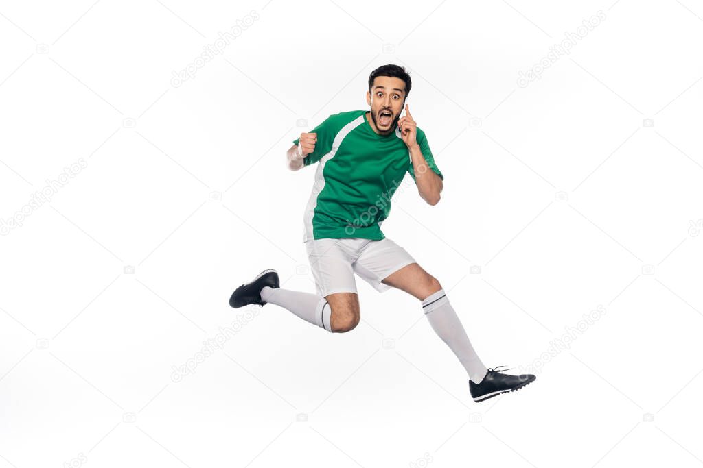 amazed football player in uniform jumping while talking on smartphone on white 