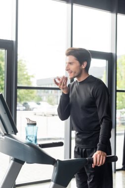 Gülümseyen adam spor salonunda koşu bandında çalışırken akıllı telefondan sesli mesaj kaydediyor. 