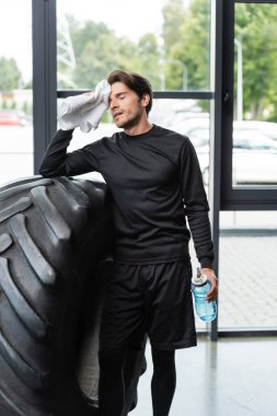 Esmer sporcu havluyla yüzünü siliyor ve spor arabasının lastiğinin yanında spor şişesi tutuyor. 
