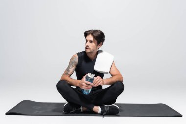 Havlusu ve spor şişesi olan esmer sporcu gri arka planda fitness minderinde oturuyor.