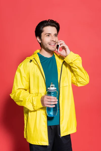 Deportista Sonriente Hablando Smartphone Sosteniendo Botella Deportiva Sobre Fondo Rojo — Foto de Stock