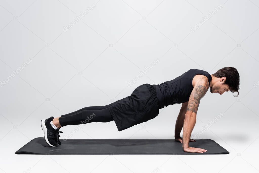 Side view of sportsman in back sportswear doing plank on fitness mat on grey background