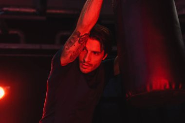 Dövmeli sporcu, Koyu renk spor merkezinde kum torbasının yanında duruyor. 