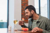 young man with closed eyes holding delicious sushi with chopsticks near glass or white wine
