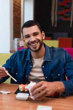 Yemek çubukları ve soya sosu kabı yanında suşi sarmalarken kameraya gülümseyen genç adam.