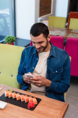 Gülümseyen adam restoranda lezzetli suşinin yanında cep telefonuyla mesajlaşıyor.