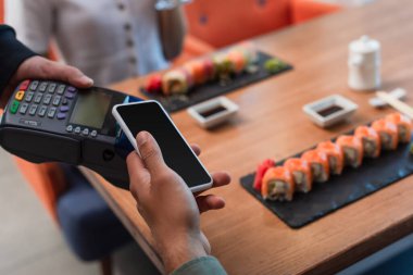 Suşi dürümleriyle servis edilen ödeme terminali ve masa başında elinde boş ekranla akıllı telefon tutan adamın kırpılmış görüntüsü.