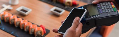 Ödeme terminali yakınında boş ekranlı akıllı telefonu olan ve bulanık suşi ruloları ve afişi olan bir adam görüntüsü.