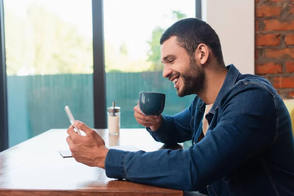 陽気な男がコーヒーカップを持ってカフェに座って携帯電話を見て — ストック写真