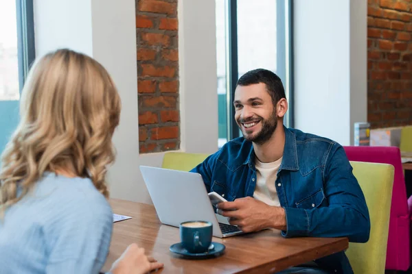 Hombre Alegre Con Teléfono Móvil Mirando Mujer Borrosa Cerca Computadora — Foto de Stock