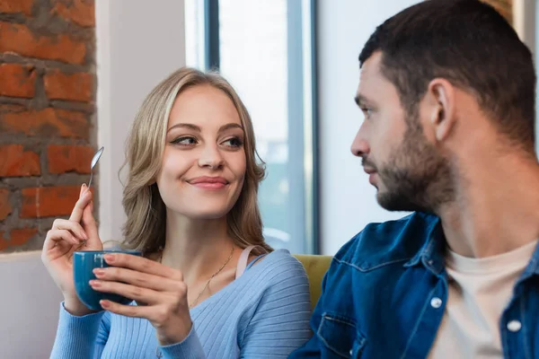 Cheerful Blonde Woman Holding Coffee Cup Teaspoon Blurred Boyfriend — Fotografia de Stock