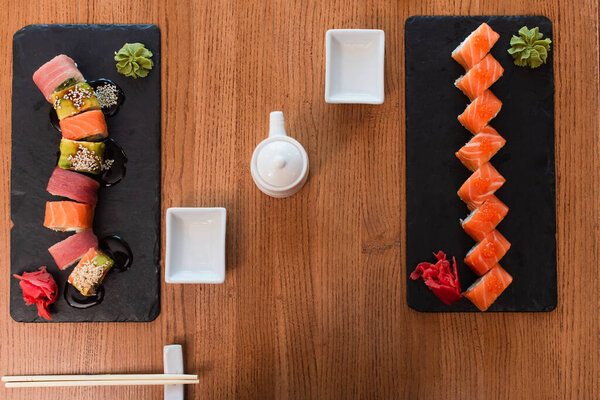 top view of black plates with delicious sushi rolls near chopsticks, soy sauce pot and bowl on wooden table