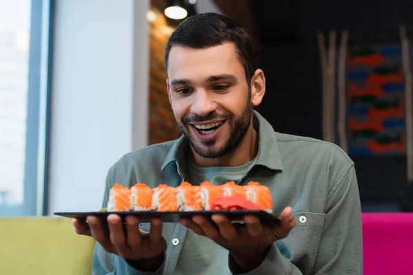 amazed man holding plate with set of tasty sushi rolls in restaurant