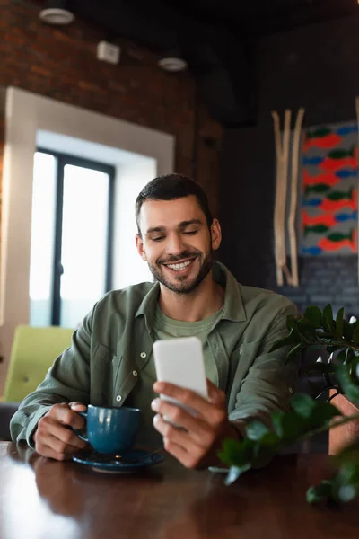 Hombre Feliz Mirando Teléfono Móvil Mientras Sostiene Taza Café Restaurante — Foto de Stock