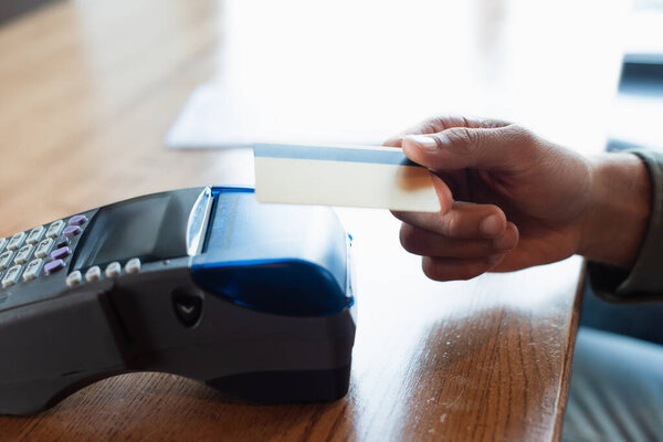 partial view of male hand with credit card near payment terminal on blurred table in cafe
