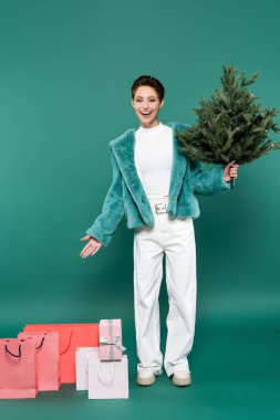 Küçük Noel ağacı olan mutlu ve modaya uygun bir kadının yeşildeki alışveriş çantalarını göstermesi.