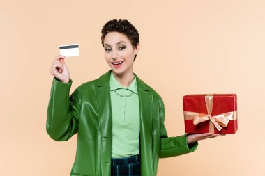 Yeşil ceketli mutlu kadın bej rengi bir kutuda kırmızı hediye kutusu ve kredi kartıyla duruyor.