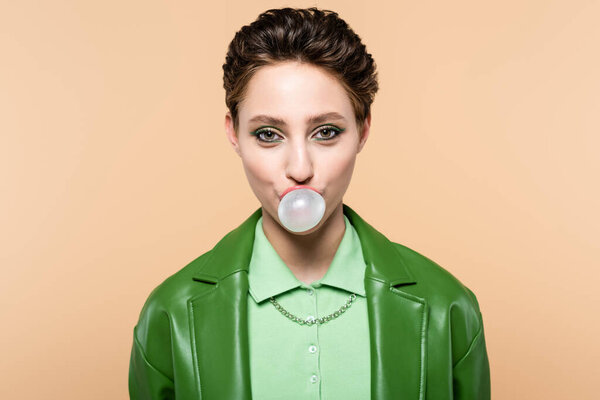 trendy brunette woman blowing bubble gum while looking at camera isolated on beige