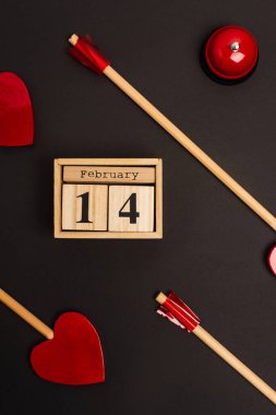 top view of heart-shaped arrows near wooden cubes with 14 february lettering and bell isolated on black  clipart