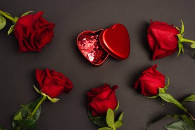 Üzerinde kırmızı konfeti kalpleri olan metalik kutunun üst görüntüsü siyah güllerin yanında.