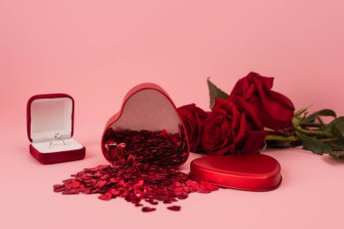 Kırmızı güllerin yanında konfeti ve pembe nişan yüzüğü olan metal kalp şeklinde bir kutu. 