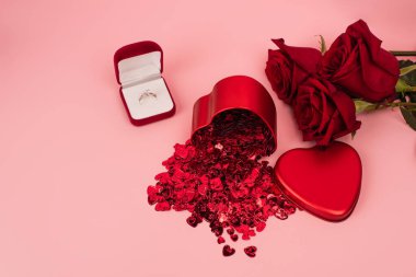 Kırmızı güllerin yanında konfeti ve pembe nişan yüzüğü olan metalik kalp şeklindeki kutunun yüksek açılı görüntüsü 
