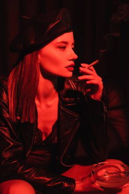 sexy woman in leather beret, jacket and bodysuit smoking in red light on black background clipart
