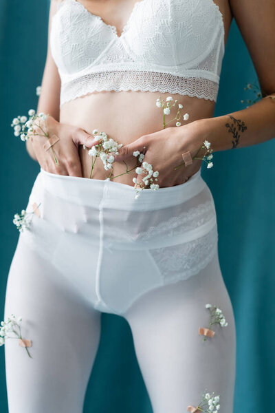 partial view of sexy woman in bra and white tights, with tiny flowers on body, posing on turquoise background