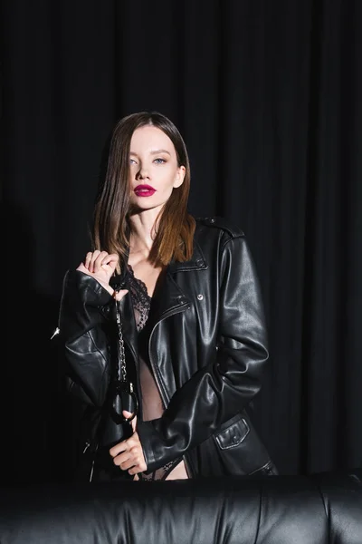 hot woman in leather jacket and lace bodysuit looking at camera while holding handcuffs on black background with drapery