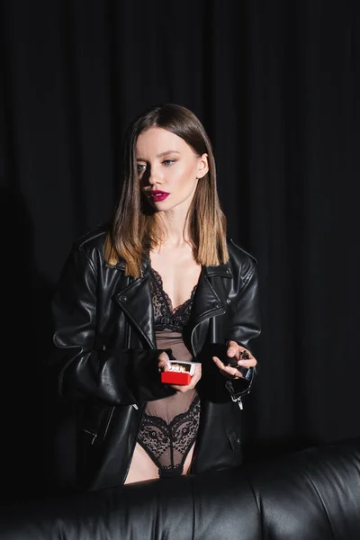 sexy woman in lace bodysuit and leather jacket holding cigarettes and lighter on black draped background