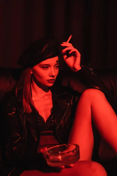 身穿贝雷帽 紧身衣和皮夹克的性感女人坐在烟灰缸和红色背景香烟上 — 图库照片