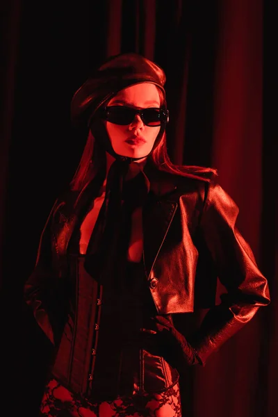 穿着皮夹克 贝雷帽和黑色太阳镜的时髦女人 双手放在臀部上 背景是深红色的 背景是深红色的 — 图库照片