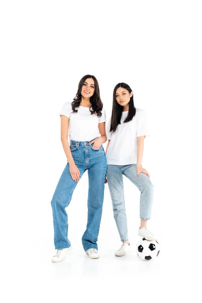 full length view of happy interracial women in jeans standing near soccer ball on white