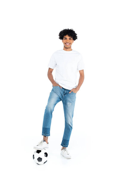 smiling african american man standing with hands in pockets of jeans near soccer ball on white