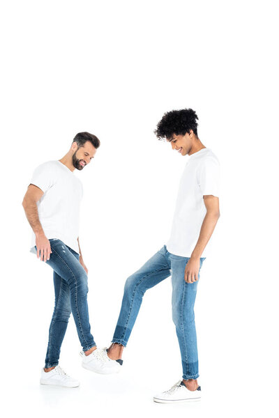 full length view of joyful multiethnic men in t-shirts and jeans having fun on white