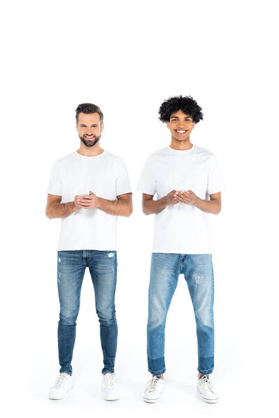 full length view of happy multiethnic men in t-shirts and jeans chatting on smartphones on white