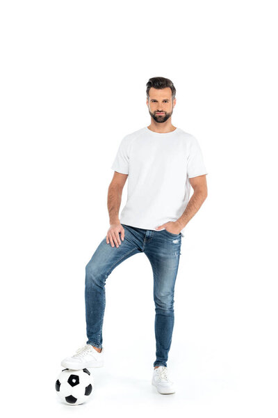 full length view of man stepping on soccer ball while standing with hand in pocket of jeans isolated on white