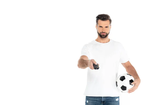 bearded man in t-shirt clicking tv channels on remote controller while holding soccer ball isolated on white
