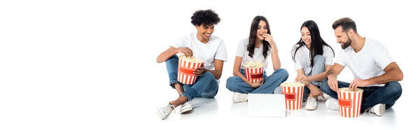 cheerful multiethnic friends watching movie on laptop and eating popcorn while sitting on white, banner