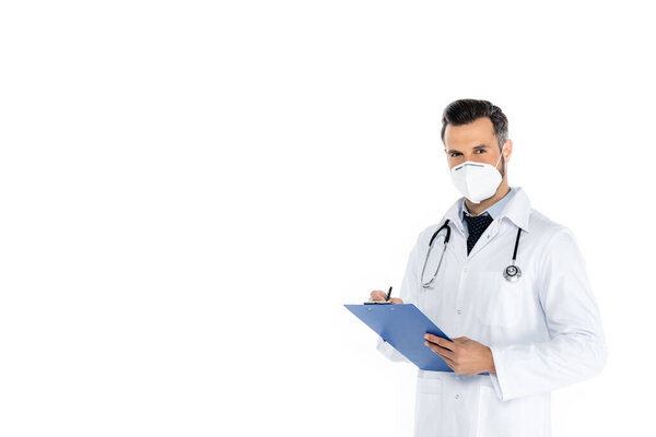 doctor in white coat and medical mask writing on clipboard and looking at camera isolated on white