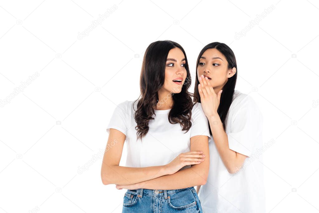 asian woman telling secret to surprised friend standing with crossed arms isolated on white