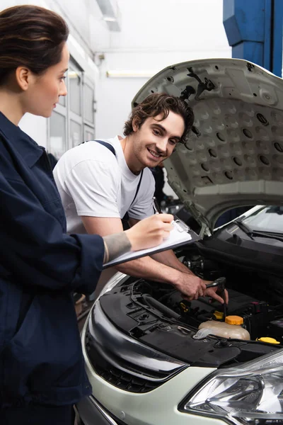Smiling Mechanic Working Car Colleague Writing Clipboard Service — Zdjęcie stockowe