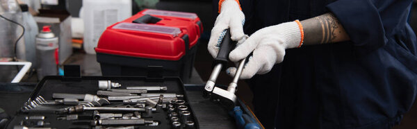 Cropped view of tattooed mechanic holding tool near toolbox in car service, banner 