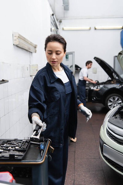 Young mechanic taking tool near auto and blurred colleague in garage 