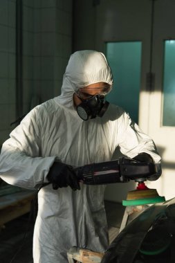 Workman in hazmat suit holding car polisher near part in garage  clipart