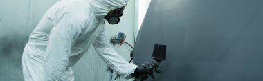Workman in hazmat suit spraying varnish on car part in garage, banner   clipart