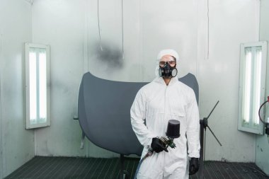 Workman in protective suit and respirator holding aerograph and looking at camera in garage  clipart
