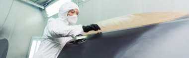 Koruyucu takım elbiseli ve maskeli bir işçinin düşük açılı görüntüsü garajda araba parçasıyla çalışıyor, afiş. 